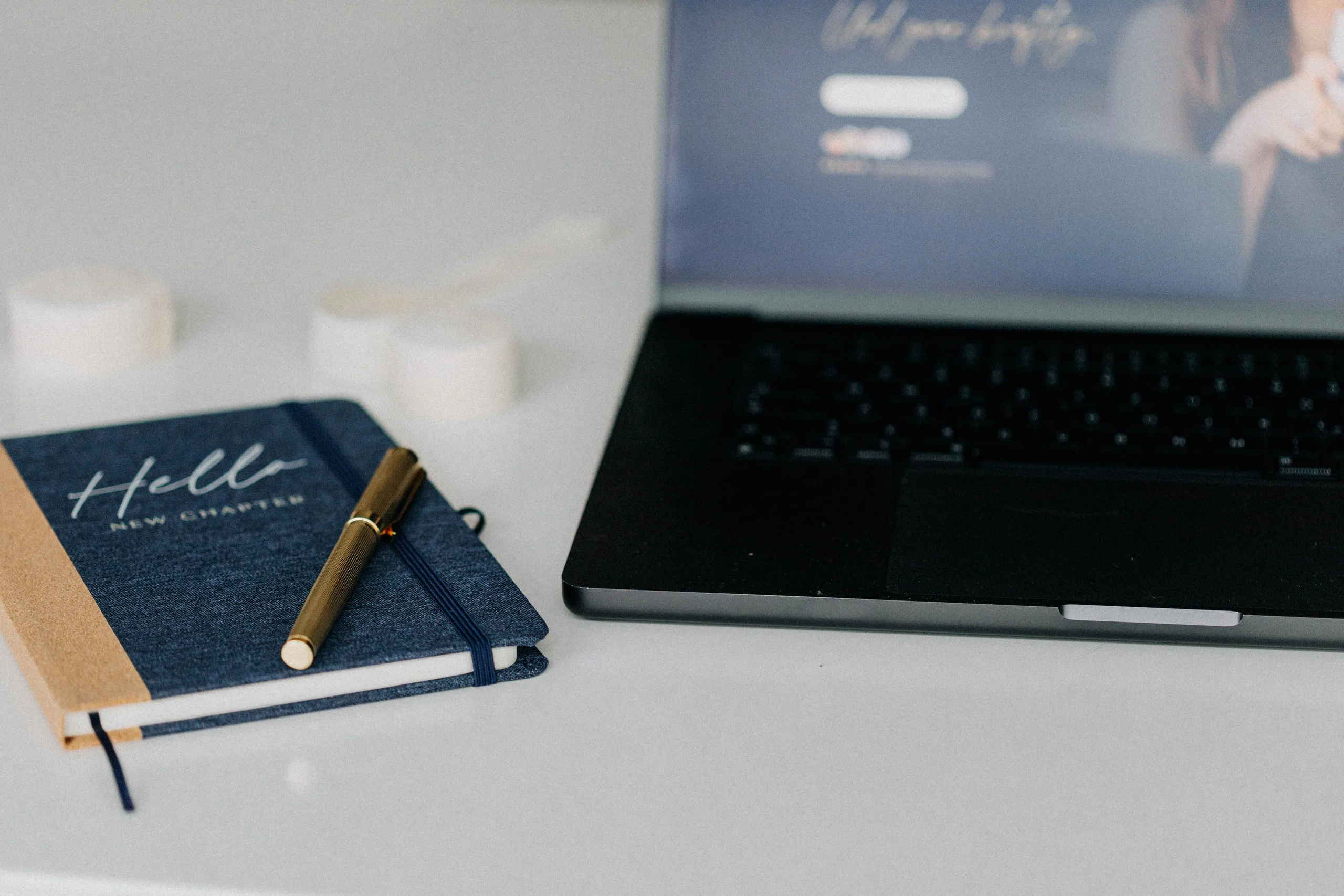 Notizbuch Hello New Chapter mit goldenem Stift neben dem Laptop, auf dem Bildschirm eine Website
