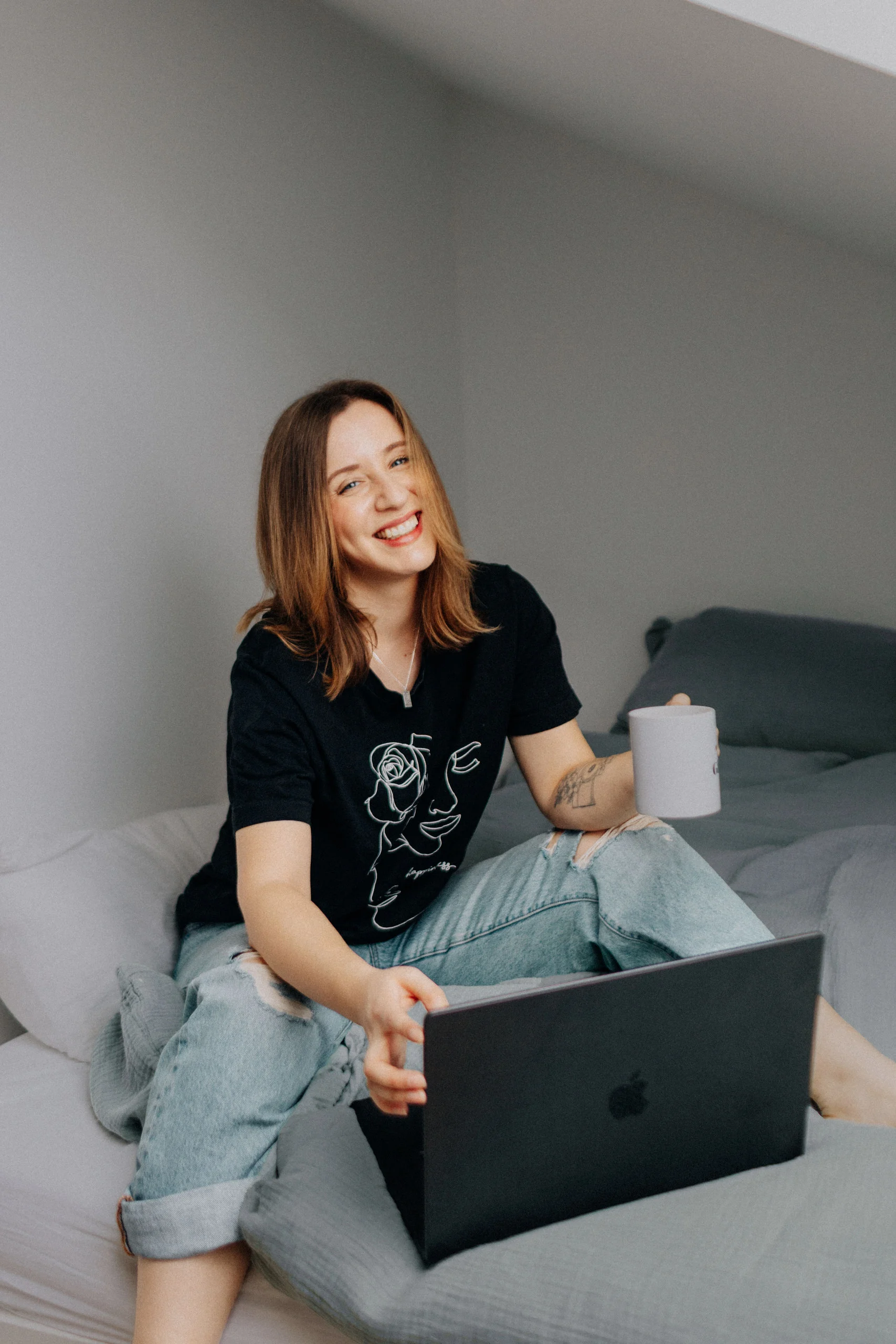 Annika Gievers sitzt mit Laptop und Kaffeetasse auf dem Bett und lacht